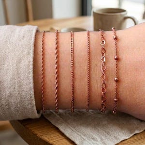 May include: Six rose gold-toned metal bracelets arranged in a row. The set includes two rope chains, one figaro chain, one simple cable chain, one chain featuring small open heart links, and one chain accented with small round beads.
