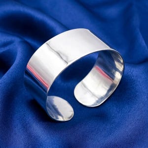 May include: A wide, polished silver cuff bracelet. The open-ended bracelet has a smooth, reflective surface. The jewelry is displayed on a vibrant blue satin fabric, creating a striking contrast. The cuff is a bold, minimalist accessory.