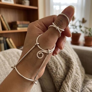 May include: A silver hand chain and rings set. The hand chain connects to a ring on the thumb and a ring on the index finger. The chain has a decorative swirl and bar design. The rings are simple bands and a knot design.