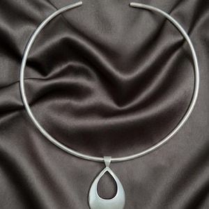 May include: A silver-colored necklace with a teardrop pendant. The necklace has a rigid, circular design that sits around the neck, with an open back. The pendant is a smooth, polished teardrop shape with a cutout center.