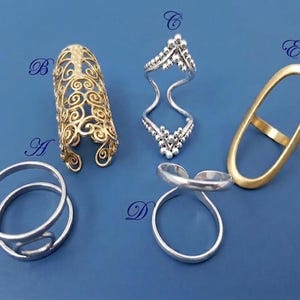 May include: Five rings labeled A through E. Ring A is a silver double-band ring. Ring B is a gold-tone filigree armor ring. Ring C is a silver chevron-shaped ring with beaded details. Ring D is a silver crossover ring. Ring E is a matte gold oval ring.