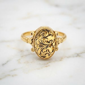 Könnte beinhalten: Ein goldfarbener Ring mit einem ovalen, floral gravierten Schmuckstein. Der Ring hat einen Band und dekorative Elemente an den Seiten des Ovals. Der Ring ist vor einem weißen Marmorhintergrund platziert. Antikes Schmuckdesign.