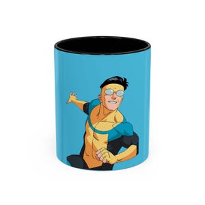 Op de afbeelding: Een keramische koffiemok met een zwart interieur en een lichtblauwe buitenkant met een cartoonillustratie van een superheld. De superheld is afgebeeld in een geel en blauw kostuum met zwarte accenten en draagt een bril.