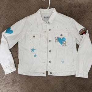 Puede incluir: Chaqueta vaquera blanca con adornos de corazón y estrella azul claro. Presenta las letras "KR" y un parche que dice "Las Vegas". La chaqueta es de Old Navy.