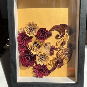 Puede incluir: Obra de arte en papel enmarcada con un diseño en forma de corazón compuesto por flores de papel burdeos, doradas y crema. La obra está sobre un fondo dorado dentro de un marco negro, creando un efecto dimensional.
