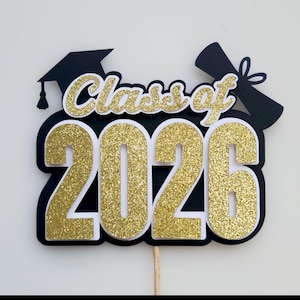 Puede incluir: Un adorno para pastel con purpurina negra y dorada que presenta el texto Class of 2026. El diseño incluye una silueta de birrete de graduación negro a la izquierda y un diploma enrollado con un lazo a la derecha, sobre un palito de madera.