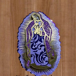 Puede incluir: Una pieza de arte religioso en capas que presenta una figura con una túnica morada con detalles dorados. La obra de arte tiene un acabado brillante y texturizado y está sobre un fondo de madera. El texto "Krafteescreations" está en la parte inferior.