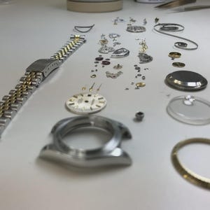 Puede incluir: Un reloj de pulsera desmontado con varios componentes esparcidos sobre una superficie blanca. Las piezas incluyen una correa de reloj plateada y dorada, una esfera, una caja, un cristal y numerosos tornillos y engranajes pequeños.