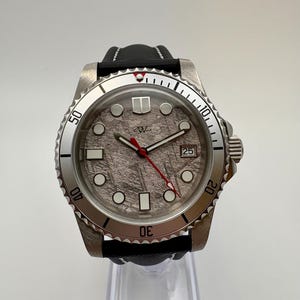 Könnte beinhalten: Eine Edelstahl-Armbanduhr mit einem strukturierten grauen Zifferblatt, einem roten Sekundenzeiger und einer Datumsanzeige bei 3 Uhr. Die Uhr hat eine drehbare Lünette und ein schwarzes Lederarmband. Das Zifferblatt hat ein Logo bei 12 Uhr.