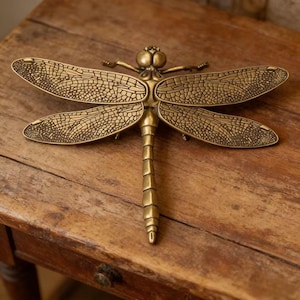 May include: A detailed brass dragonfly sculpture with textured wings and body. The dragonfly is a golden color and is resting on a wooden surface. The wings are spread wide, showcasing intricate vein patterns.
