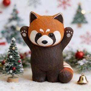 Puede incluir: Figura de madera de panda rojo con los brazos levantados. El panda tiene la cabeza y el cuerpo de color marrón rojizo, con detalles blancos alrededor de los ojos y la boca. La figura está sobre un fondo nevado con adornos navideños.