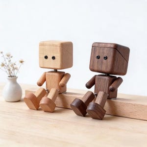 Pode incluir: Duas figuras de robôs de madeira, uma castanho-claro e outra castanho-escuro, estão lado a lado. Cada uma tem uma cabeça em forma de cubo com olhos de pontos pretos, braços pequenos e pernas. Estão posadas como se estivessem sentadas, com as pernas estendidas.