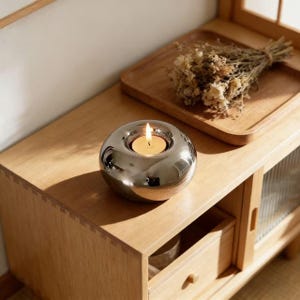 May include: A polished silver candle holder with a lit tea light candle. The spherical holder sits on a wooden surface, with a wooden tray and dried flowers in the background. The candle flame glows brightly.