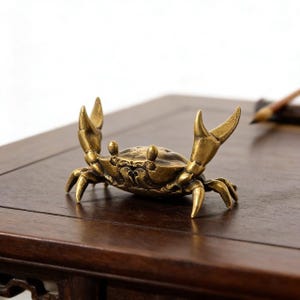 May include: A detailed brass crab figurine with raised claws and intricate detailing. The crab is a golden color and sits on a dark wooden surface. This decorative item could be used as a paperweight or displayed as a unique accent piece.