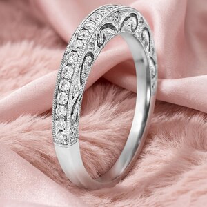Könnte beinhalten: Ein silberner Ring mit geschwungenem Design, der eine Reihe kleiner, klarer Steine und aufwendige Filigranarbeiten aufweist. Der Ring ist vor einem weichen rosa Hintergrund platziert, der seine eleganten und zarten Merkmale hervorhebt. Der Ring ist ein Schmuckstück.