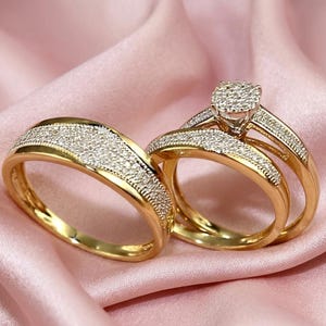 May include: A set of gold rings, one with a central cluster of small, clear stones and two bands, and another with a single band. The rings are set against a pink satin background.