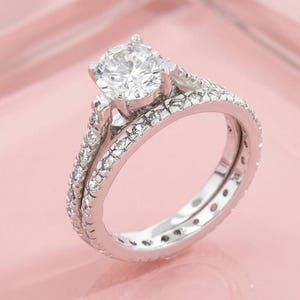 Könnte beinhalten: Ein silberfarbenes Verlobungsring-Set mit einem großen, runden, im Brillantschliff geschliffenen Mittelstein in einer Vier-Krappen-Fassung. Sowohl der Ring als auch der passende Ehering sind außen mit kleinen, klaren, rund geschliffenen Steinen besetzt.