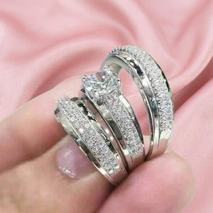 May include: A set of three silver-toned rings featuring a central ring with a large round-cut clear stone and two flanking bands. Each band is encrusted with numerous small, sparkling clear stones that cover the entire visible surface area.