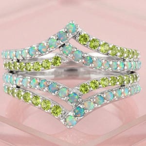 May include: A silver chevron ring featuring two bands set with alternating round-cut opals and lime green peridot gemstones. The design creates a V-shaped silhouette with a double-row arrangement of small, faceted stones embedded in the metal.
