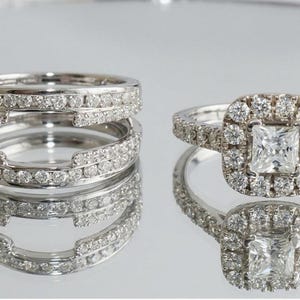 Puede incluir: Dos anillos de plata con pequeñas gemas transparentes. Un anillo tiene un halo de gemas alrededor de una piedra central cuadrada. El otro anillo tiene un diseño de doble banda con gemas. Los anillos están sobre una superficie reflectante.