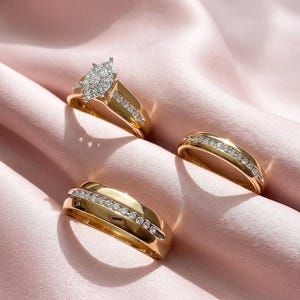 Può includere: Tre anelli d'oro con dettagli di diamanti sono esposti su una superficie di raso rosa. Un anello presenta un gruppo di diamanti a forma di pera, mentre gli altri due hanno una fila di diamanti più piccoli. Gli anelli sono probabilmente per un matrimonio o un fidanzamento.
