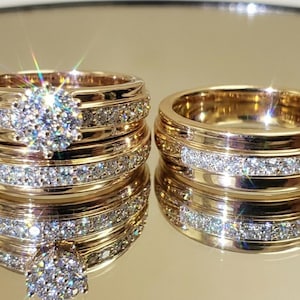 Pode incluir: Dois anéis dourados com detalhes brilhantes de diamantes. Um anel apresenta um grande diamante central rodeado por pedras menores, o outro uma faixa de diamantes. Os anéis são exibidos em uma superfície reflexiva.