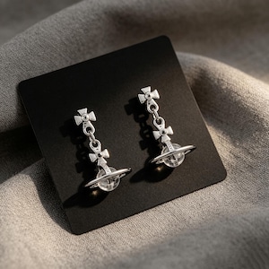 Op de afbeelding: Een paar zilverkleurige oorhangers. Elke oorbel heeft een kruisvormige bovenkant, een ketting en een transparante bol met een zilveren ring. De oorbellen worden gepresenteerd op een zwarte kaart tegen een grijze stoffen achtergrond.