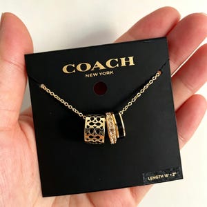 May include: Gold-coloured necklace with three pendant rings. One ring has a cut-out pattern, another is encrusted with small crystals, and the third is black. The necklace is on a black card with the brand name "COACH NEW YORK".