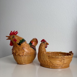 May include: Two woven wicker baskets shaped like chickens. The left basket is a closed container with a rooster head, red comb, and green and black accents. The right basket is an open, shallow bowl with a smaller chicken head and tail detail.