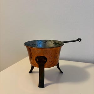 Op de afbeelding: Een kleine koperen pan met een metalen binnenkant, voorzien van drie donkere metalen poten en een lange, slanke handgreep van donker metaal. De buitenkant van de pan heeft een warme, gepolijste koperen afwerking met zichtbare ouderdomssporen.