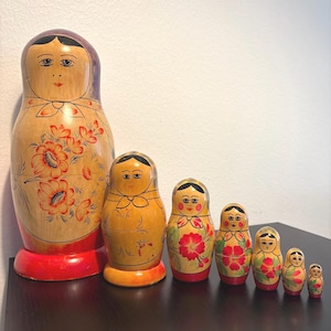 Könnte beinhalten: Ein Set aus sieben hölzernen Matroschka-Puppen, die in einer Reihe von der größten zur kleinsten angeordnet sind. Jede Puppe hat ein handbemaltes Gesicht mit schwarzem Haar, ein rotes und oranges Blumenmuster und einen roten oder orangen Sockel.
