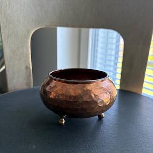 Peut inclure: Un petit bol en cuivre martelé avec un intérieur sombre et trois petits pieds en laiton. Le bol a une forme arrondie et une surface texturée, reflétant la lumière. Il repose sur une surface sombre.