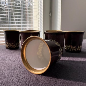May include: A set of six dark brown ceramic pots with gold rims and floral designs. One pot is lying on its side, revealing a white interior. The pots are arranged on a purple surface near a window.