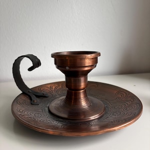 May include: A copper-colored metal candle holder featuring a circular base with an embossed floral pattern. The center holds a cylindrical candle cup, and a curved, textured black metal handle is attached to the side of the base.