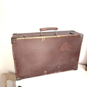 May include: A rectangular brown suitcase with a textured surface, featuring a wooden top handle, metal corner reinforcements, and metal latches. The exterior shows signs of wear, including scuffs and a faded marking on the front panel.
