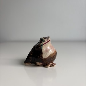 Op de afbeelding: Een klein, met de hand gevormd keramieken kikkerbeeldje met een glanzende afwerking. Het object heeft een rond lichaam met een kleurenpalet van donkerbruin, crème en accenten van groen. De kikker zit in een zittende houding en kijkt met een open mond omhoog.