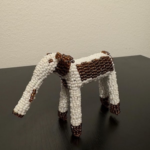 May include: A beaded elephant ornament. The elephant is white with brown detailing on its back, trunk, and feet. The figurine is made of small beads and has a detailed design. The elephant is standing on four legs and has a long trunk.