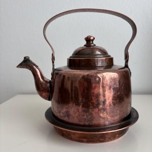 Op de afbeelding: Een gepolijste koperen theepot met een rond lichaam, een gebogen tuit en een hoog gebogen handvat. De theepot heeft een bijpassend deksel met een kleine knop en staat op een ronde koperen voet, met een warme, metalen afwerking.