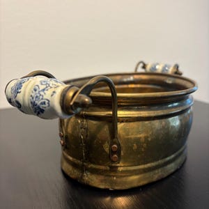 May include: An antique brass oval-shaped container with two handles. The handles are attached to the container with rivets. The handles have white ceramic grips with blue floral patterns. The container is on a dark surface.