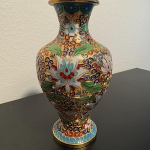 Pode incluir: Um vaso decorativo cloisonné com borda e base douradas. O vaso apresenta um design floral detalhado em branco, vermelho, azul e verde sobre um fundo dourado. O vaso tem aproximadamente 20 cm de altura.