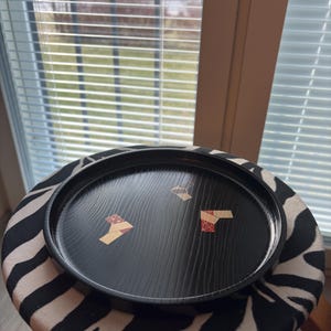 May include: A round, black wooden tray with a decorative grain pattern and inlaid geometric designs. The tray is placed on a zebra-print stool. The tray's diameter is approximately 12 inches.