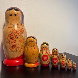 May include: A set of seven wooden Matryoshka dolls, also known as Russian nesting dolls. The dolls are painted with floral designs in red and orange on a light brown background. The largest doll is about 10 inches tall.