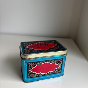 Peut inclure: Une boîte à thé rectangulaire en métal avec un fond bleu vif, des panneaux noirs et des cartouches décoratives rouges. Le couvercle et la face avant présentent des textes en cyrillique, dont "ЧАЙ ГРУЗИНСКИЙ ЭКСТРА" et "МПП РСФСР РОСДИЕТЧАЙПРОМ".