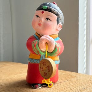 Op de afbeelding: Een keramieken beeldje van een kind in traditionele Chinese kledij, met een houten munt. Het figuur draagt een rode jurk met gouden en blauwe accenten, een zwarte hoed en heeft een geschilderd gezicht. De munt heeft Chinese karakters.