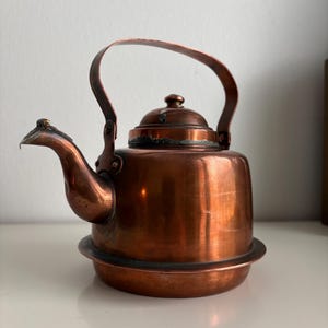 Peut inclure: Une bouilloire à thé en cuivre poli avec un corps arrondi, un bec verseur incurvé et une anse fixe arquée. La bouilloire comprend un couvercle amovible avec un petit bouton, présentant des signes de patine naturelle et d'usure sur les bords.
