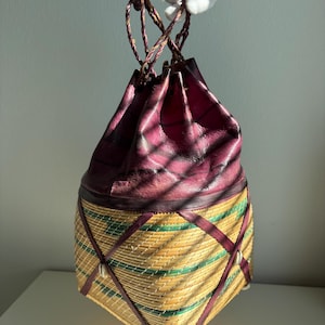 May include: A drawstring bag featuring a deep purple leather top section with braided leather handles and a woven straw base in natural tan and green. The straw body is accented with thin purple leather strips in a crisscross pattern and small white shells.