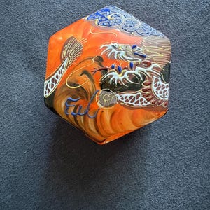 Könnte beinhalten: Sechseckige orangefarbene und braune dekorative Box mit einem detaillierten Drachendesign. Die Box hat ein blaues und weißes Wolkenmuster am oberen Rand. Der Drache ist mit komplizierten Details und einem goldenen Akzent dargestellt.