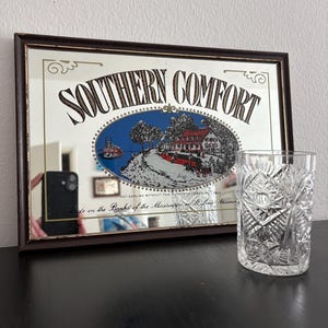 Peut inclure: Un miroir rectangulaire encadré de marron, avec l'inscription "SOUTHERN COMFORT". Il présente une illustration d'un bateau fluvial et d'une maison. Un verre clair et décoré est placé à droite.