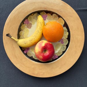 May include: A yellow banana, a red apple, and an orange rest on a decorative plate. The plate has a light brown rim and a floral design in the center. The background is a dark gray surface.