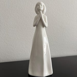 Könnte beinhalten: Eine weiße Keramikfigur einer Person mit gefalteten Händen im Gebet. Die Figur trägt ein langes Kleid und hat kurzes Haar. Die Figur steht auf einer schwarzen Oberfläche.